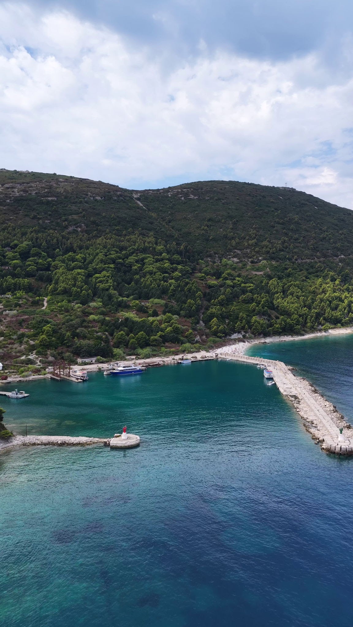HD aerial drone image showing the port of Sazan Island, with the harbour, blue water and surrounding coastline illuminated by the sun. The image captures the island's coastal infrastructure, clear sea and scenic Mediterranean landscape.
